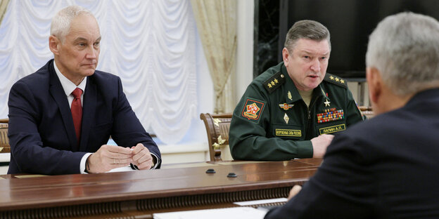 Andrei Belousov und Alexander Lapin sitzen zusammen an einem Tisch, ihnen gegenüber steht ein Mann, der nicht wiederzuerkennen ist – sie sehen ernst aus