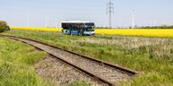 Zugschienen, ein Bus und im Hintergrund ein gelbblühendes Feld mit WIndrädern und Stromleitungen.