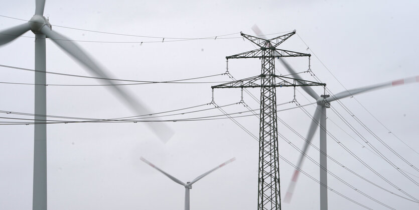 Windräder drehen sich hinter Stromleitungen bei grauem Himmel