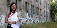 Eine junge Frau steht vor einem verlassenen Gebäude, drum herum lange Gräser.