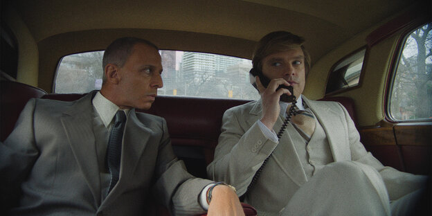 Two men in suits in the backseat of a car, one on the phone