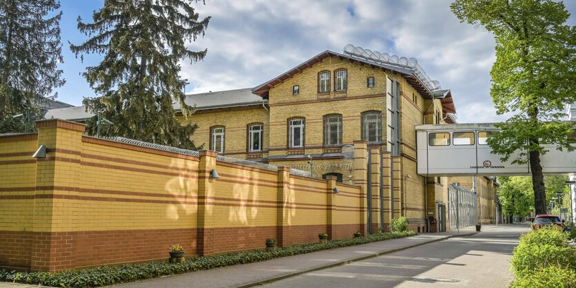 Ein Backsteingebäude ist von einer hohen Mauer und Stacheldraht umgeben, man sieht außerdem eine völlig umschlossene Brücke, die von einem Gebäude ins nächste führt.