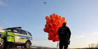 Ein Polizist steht auf einem Flugfeld und hält viele rote Luftballons, ein anderer steht in Warnweste neben einem Polizeiauto.