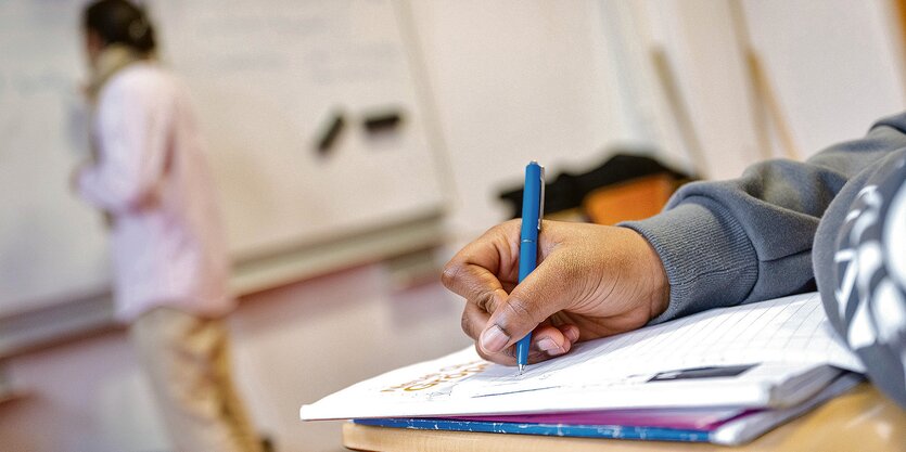 Eine Kinderhand hält einen Stift und schreibt in ein Heft, im Hintergrund eine Person an der Tafel