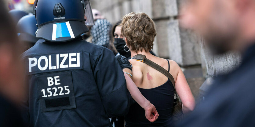 EIn Polizist mit Helm führt eine junge Frau ab. Beide sind von hinten zu sehen.