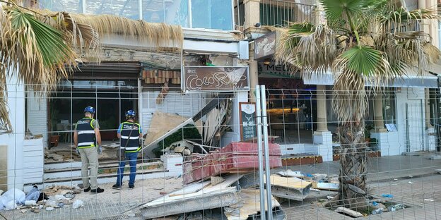 Zwei Polizisten stehen vor einer umzäunten Bar. Vor ihnen sind Trümmer zu sehen. Neben ihnen steht eine Palme.