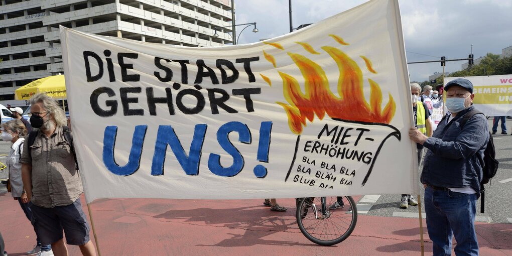 Szene auf einer Demonstration am 11. 09. 2021 am Alexanderplatz unter dem Motto "Wohnen für Alle - Gemeinsam gegen hohe Mieten und Verdrängung": Zwei Männer halten ein Plakat mit der Aufschrift "Die Stadt gehört uns!" Daneben abgebildet: Eine brennende Mieterhöhung.