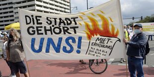 Szene auf einer Demonstration am 11. 09. 2021 am Alexanderplatz unter dem Motto "Wohnen für Alle - Gemeinsam gegen hohe Mieten und Verdrängung": Zwei Männer halten ein Plakat mit der Aufschrift "Die Stadt gehört uns!" Daneben abgebildet: Eine brennende Mieterhöhung.