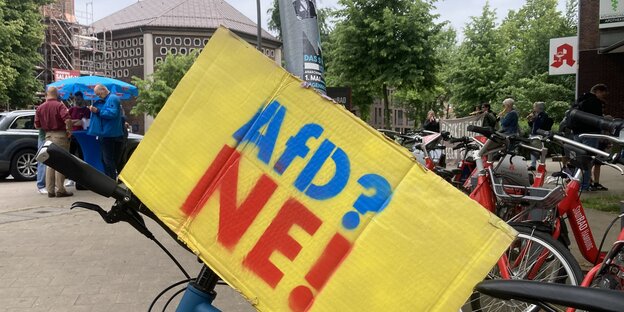 Im Vordergrund ein abgestelltes Fahrad mit der Aufschrift "AfD? Nee!". Dahinter ein AfD-Wahlkampfstand (links) und blockflötengestützter Protest (rechts)