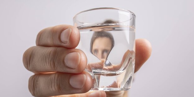 Eine Hand hält ein Wasserglas, ein gesicht spiegelt sich darin