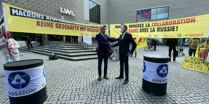 Demonstration mit Transparenten, Aufschrift "Keine Geschäfte mit Rosatom", zwei anzugtragende Demonstranten mit Macron- und Putin-Masken reichen sich die Hände