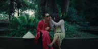 Szene in einem Park in Saigon. Zwei Menschen sitzen auf einer Mauer vor einem Baum. Die Person rechts trägt ein rotes Seidenkleid und eine Sonnenbrille. Ihren Sonnenhut hat sie neben sich abgelegt. Die Person links trägt ein gelbes Kleid und einen hellen