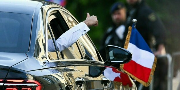 Aus seiner Staatskarosse hält der französische Präsident Macron seinen Arm und zeigt mit dem Daumen nach oben.