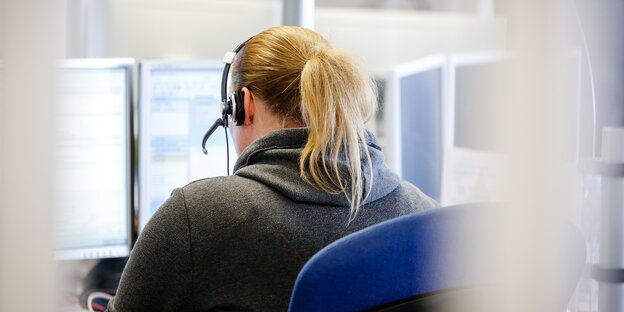Eine Frau hat Kopfhörer auf und sitzt mit dem Rücken zum Betrachter auf einem Bürostuhl und arbeitet an einem Bildschirm