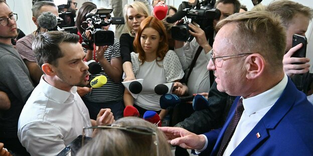 Zwei Männer umringt von Vertreter:innen der Presse sprechen intensiv miteinander.