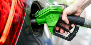 Eine Frau hält an einer Tankstelle an einer Zapfsäule eine Zapfpistole in der Hand und betankt ein Auto.