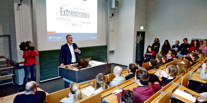 Ein Mann spricht in einem Hörsaal zu Menschen, hinter ihm leutet das Wort "Extremismus".