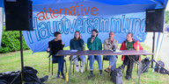 5 Menschen sitzen am Tisch unter einem blauen Zeltdach und einem Transparent "Alternative Hauptversammlung"