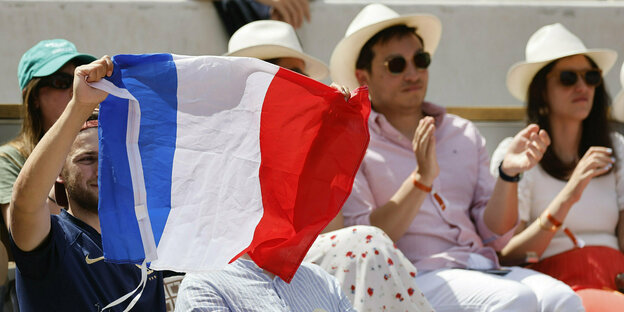 Franzosen mit Sonnenhüten sitzen auf den Rängen bei einem French Open Match, einer hält eine Trikolore hoch