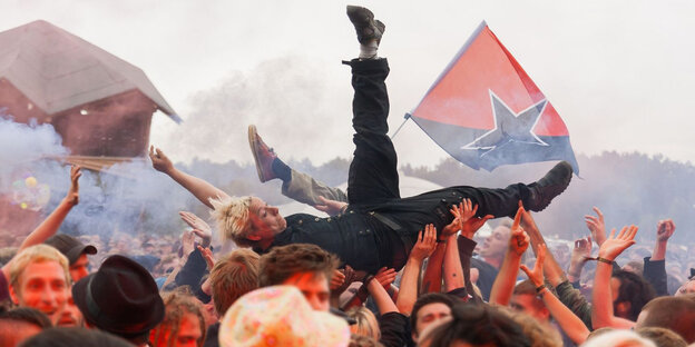 Eine Person wird auf den Händen von Festivalbesuchern gehalten, im Hintergrund eine Anarcho-Flagge