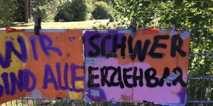 Zwei bunte Pappen mit dem Text "Wir sind alle schwer erziehbar" hängen an einem Zaun vor einer grünen Wiese.