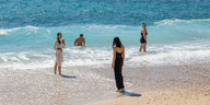 Touristinnen stehen mit den Beinen im Wasser und fotografieren, ein Mann badet im Meer