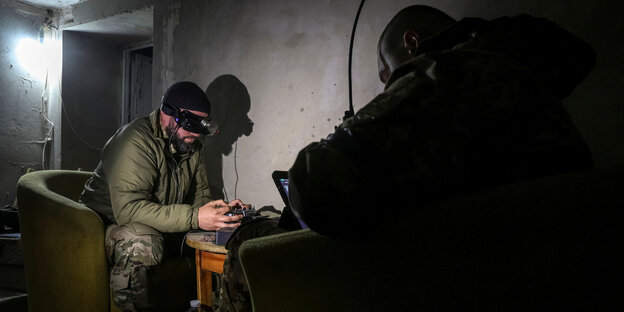 Ein ukrainischer Soldat sitzt in einem spärlich beleuchteten Raum in einem staubigen Sessel und beschäftigt sich mit einer Drohne