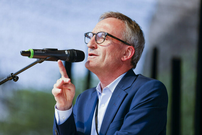 Thomas Geisel steht in blauem Anzug und offenem Hemd an einem Mikrofon und spricht mit erhobenem Zeigefinger