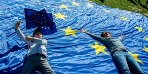 Zwei Jugendliche liegen auf einer sehr großen EU-Flagge, die auf einer Wiese ausgebreitet wurde. Der Junge hält zwei kleine EU-Flaggen in der Hand