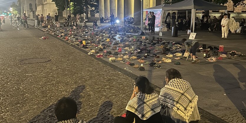 Menschen sitzen und stehen am Berliner Boulevard Unter den Linden, Kinderschuhe stehen aufgereiht, ein Redner liest am Mikrofon von einem Blatt ab