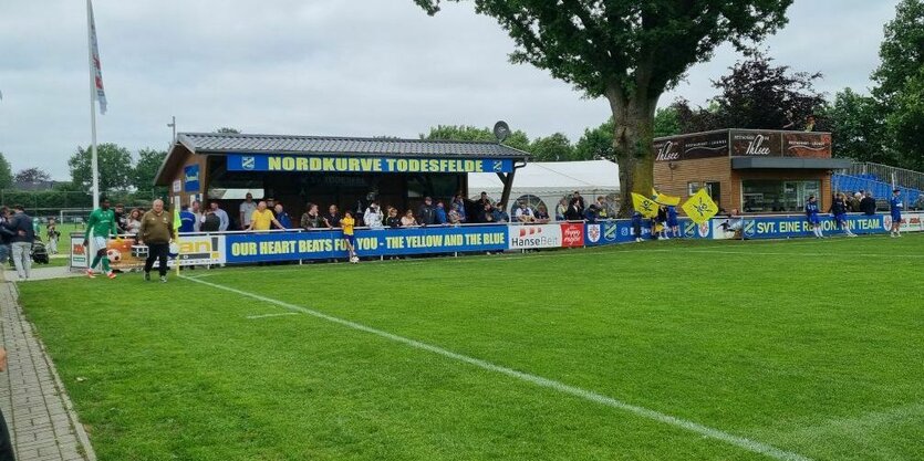 Ein Fußballfeld mit einer Tribüne auf der Nordkurve Todesfelde steht, Fußballspieler stehen angelehnt an der Absperrung davor