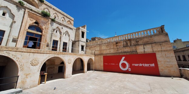 Am Alman Karargah, einem sonnenüberfluteten Gebäude aus hellem Stein mit Rundbögen, ist ein Banner der 6. Mardin Biennale angebracht.