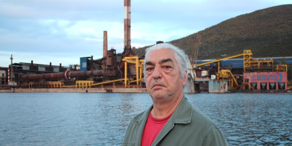 ein grauhaariger Mann mit dicken Tränesäcken steht vor einer Fabrik
