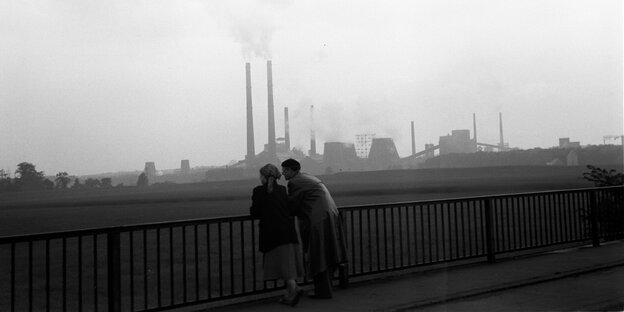 Schwarz-Weißer Filmstill, der zwei Menschen auf einer Brücke zeigt. Sie lehnen am Geländer und schauen in Richtung zweier Farbrikschlote am Horizont, aus denen Rauschschwaden aufsteigen