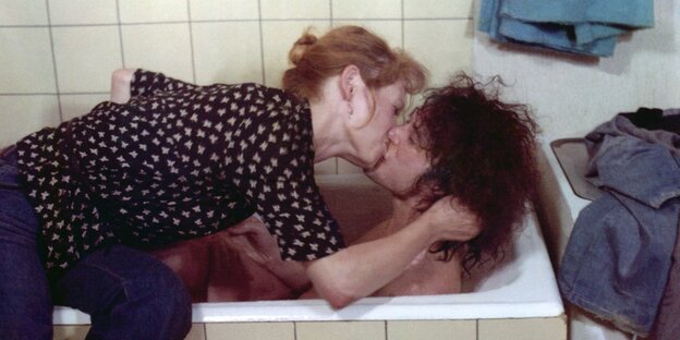 Ein Mann liegt nackt in der Badewanne in einem beige gekachelten Badezimmer. Eine Frau mit roten Haaren sitzt in Jeans und Hemd auf dem Badewannenrand, sie beugt sich zu dem Mann, fast in sein Haar und küsst ihn. Rechts steht eine Waschmaschine, auf der K