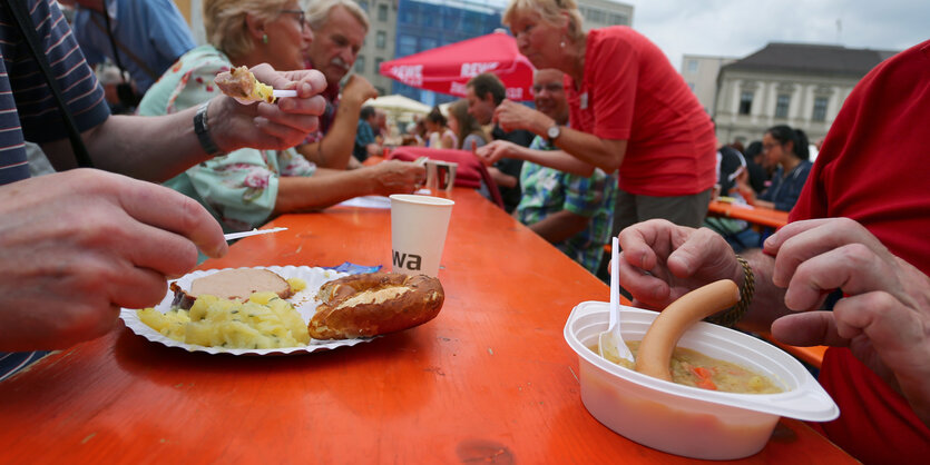 Menschen sitzen auf einem öffentlichen Platz an einem langen Biertisch, vor sich Schalen mit Bockwurst