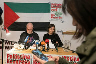 Zwei Personen sitzen an einem Tisch mit Mikrofonen, im Hintergrund hängt eine Palästina-Flagge.