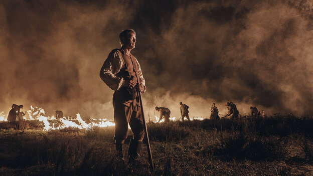 Kahlen, gespielt von Mads Mikkelsen, steht mit einer Spitzhacke vor einem brennenden Feld, das Arbeiter umpflügen, um es urbar zu machen