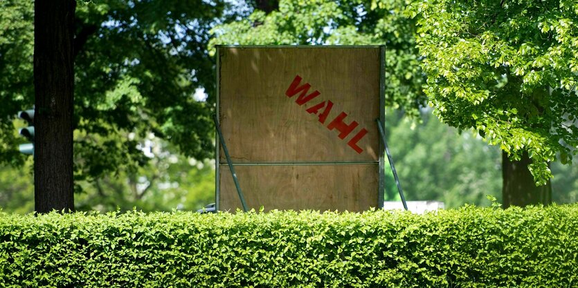 Hinter einer Hecke steht ein Wahlplakat mit der Aufschrift Wahl von hinten zwischen grünen Bäumen