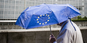 Ein Mensch geht zu Fuß vor der Europäischen Kommission entlang und trägt einen Regenschirm mit dem Symbol der Europäischen Union