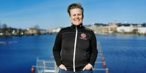 Porträt von Sara Nylund, sie steht vor einem blauen See und lacht in die Kamera. Sie lacht und trägt eine dunkle Jacke mit dem Emblem einer rote Rose, dem Logo der Sozialdemokraten