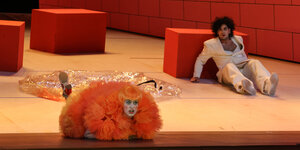 Eine Frau in orangem Kostüm liegt vorne am Bühnenrand, etwas weiter im Hintergrund liegt der ratlose Prinz