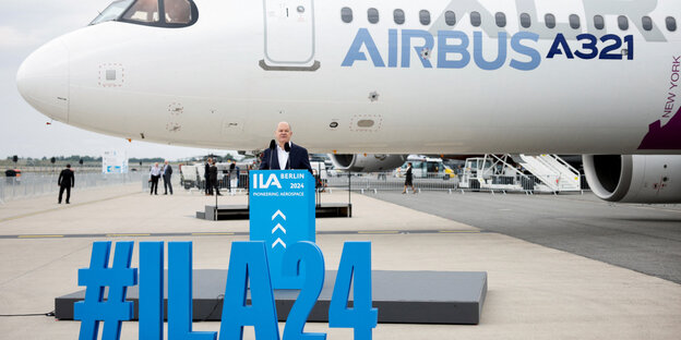 Olaf Scholz an einem Rednerpult auf einem Rollfeld, hinter ihm eine Airbus-Passagiermaschine, vor ihm ein Modell aus den Schriftzeichen #ILA24
