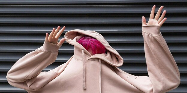 Frau mit pinken Haaren hat Kopf im rosa Hoodie versteckt, Arme sind nach oben gereckt