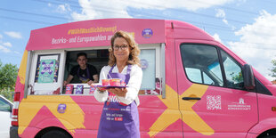 eine Frau in violetter Schürze trägt ein Tablett mit Eisbehern vor einem pinken Eiswagen mit Werbung fürs Wählen