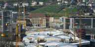 Blick auf eine Baustelle mit Kränen und weißen Planenüberdachungen, im Hintergrund ein Weinhang und Wohnhäuser