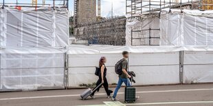 Blick auf eine Baustelle mit Kränen und weißen Planenüberdachungen, im Hintergrund ein Weinhang und Wohnhäuser