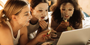 3 Mädchen liegen auf einem Bett, sie schauen in ein Labtop , tragen Gesichtsmasken , einige haben Gurkenscheiben in der Hand