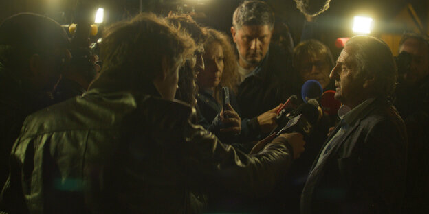 François Schaar (Daniel Auteil) steht umringt von Journalisten mit Mikrofonen.