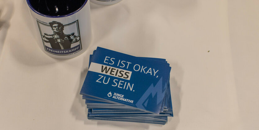 ein blauer Flyer mit der Aufschrift "Es ist okay weiss zu sein" der Jungen Alternative neben einer Tasse, Motiv Mann mit Schwert Freiheitskampf Aufschrift